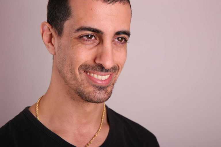 Close-up of a smiling man with facial hair and gold chain in studio setting.