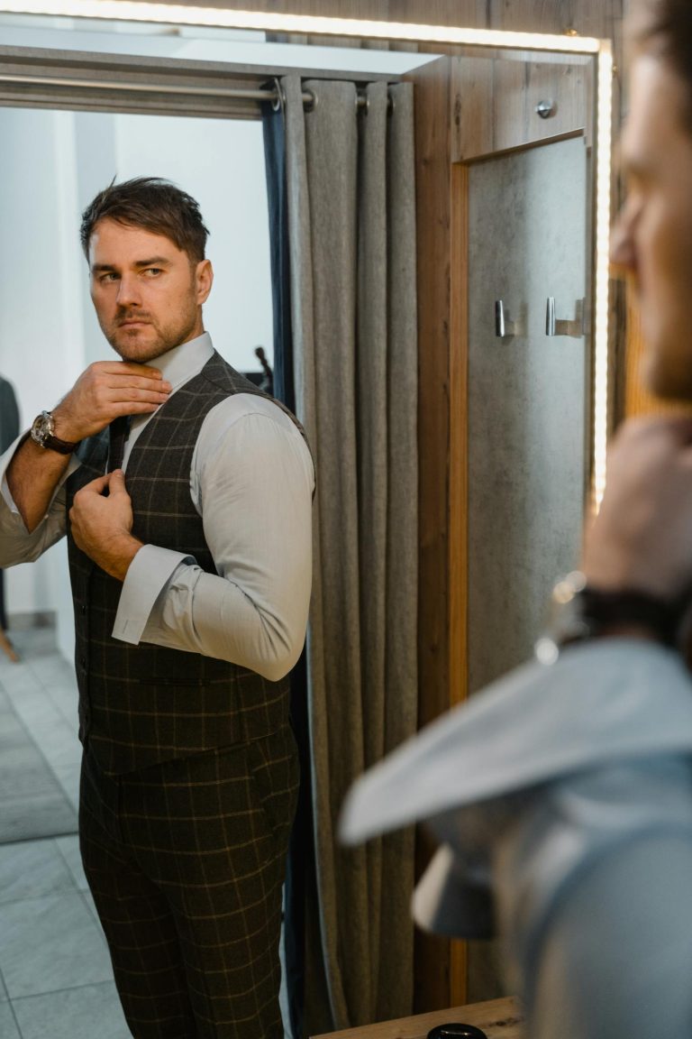 Fashionable man adjusting vest in front of a mirror, showcasing a modern and stylish outfit indoors.